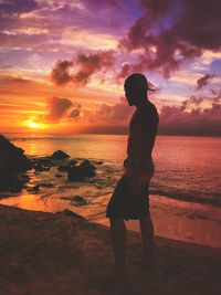 Shirtless man looking away while standing at beach against sky during sunset