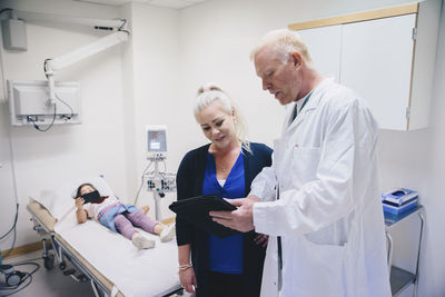 Mature doctor showing digital tablet to woman standing by bed at hospital