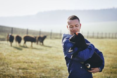 Man holding sheep while standing on field against sky