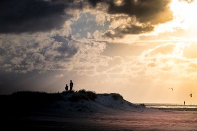 Silhouette children standing at beach against cloudy sky during sunset