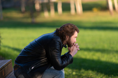 Pensive serious young bearded man wear black leather jacket sitting and thinking outdoors