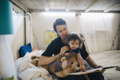 Father reading book with daughter while sitting in bedroom