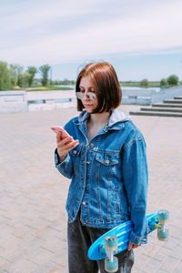 Young stylish woman walks on street with a skateboard and uses a smartphone