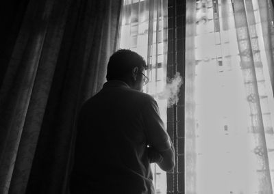 Rear view of man smoking while standing by window