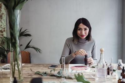 Portrait of smiling young woman preparing perfume at table against wall in workshop