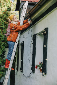 Woman decorating house with lights standing on ladder