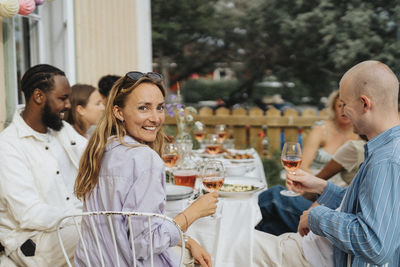 Portrait of smiling young woman looking over shoulder while sitting with friends during dinner party at cafe