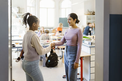 Schoolgirl doing handshake with female teacher standing in classroom