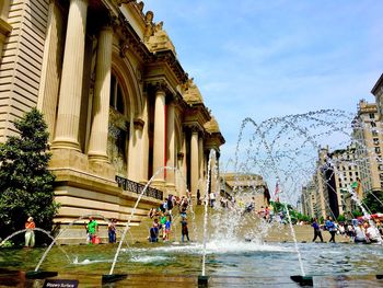 People walking on fountain in city against sky