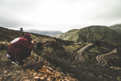 Side view of hiker photographing road while crouching on mountain against sky