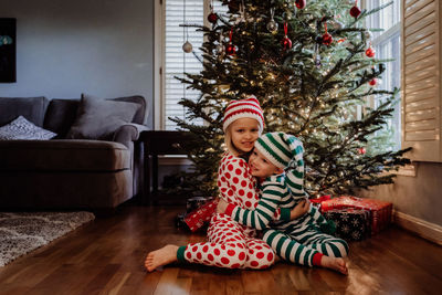 Siblings in costume sitting against christmas tree at home