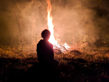 Silhouette boy sitting by campfire on field at night