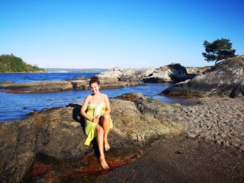 Portrait of smiling woman sitting on rock at beach against sky during summer