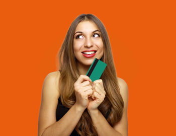 Happy young woman holding credit card while looking away against orange background
