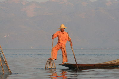 Full length of fisherman standing on fishing boat in lake against mountain