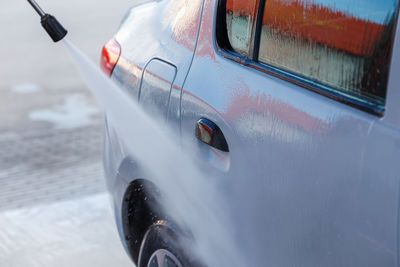 Hand with high pressure washer washing white car at public self service car washing station