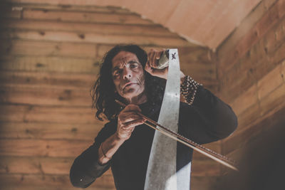 Portrait of man holding musical instrument and work tool against wooden wall