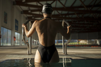 Young beautiful woman leaves the indoor pool by the stairs with black swimsuit, rear view