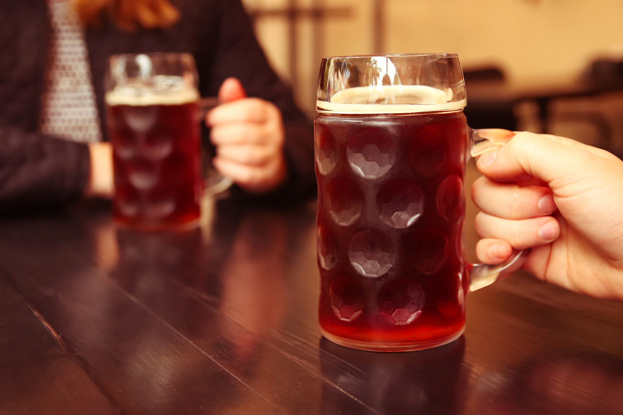 drink, refreshment, glass, alcohol, food and drink, drinking glass, bar, beer, household equipment, hand, adult, beer glass, drinking, happy hour, table, celebratory toast, alcoholic beverage, indoors, friendship, pub, soft drink, bar counter, pint glass, holding, smiling, women, red, two people, distilled beverage, celebration, men, relaxation, cold temperature, focus on foreground, togetherness, lifestyles, leisure activity, restaurant, wood, after work, frothy drink, sitting