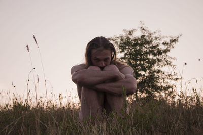 Man hugging knees on field against sky