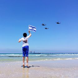 Rear view of boy jumping with israeli flag while looking at military helicopters flying over beach