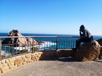 Man sitting on rock with sea in background against clear blue sky