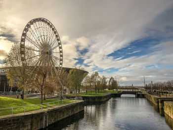 View of ferris wheel by river against cloudy sky