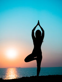 Silhouette woman doing tree pose at beach against sky during sunset