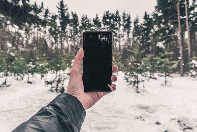 Close-up of hand holding smart phone against snow covered trees