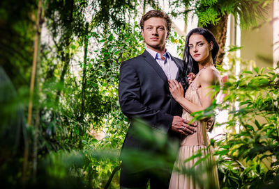 Portrait of confident bride and groom while standing amidst plants