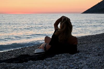 Woman sitting on shore at beach against sky during sunset