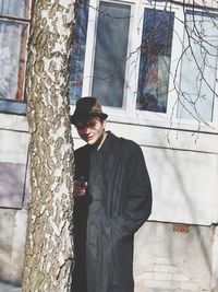 Young man wearing hat and overcoat while standing by tree trunk in city
