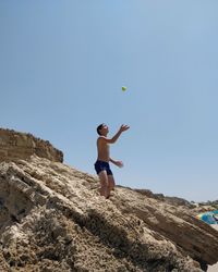 Low angle view of shirtless boy catching ball on rocky shore against clear blue sky