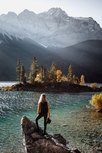 Rear view of woman standing on rock at lakeshore against mountains