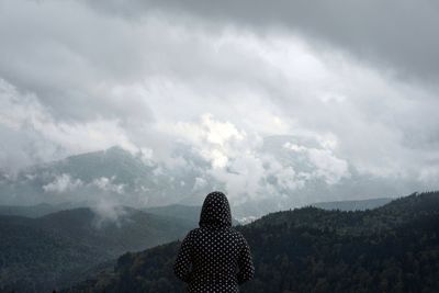 Rear view of mid adult woman looking at mountain range while standing against cloudy sky