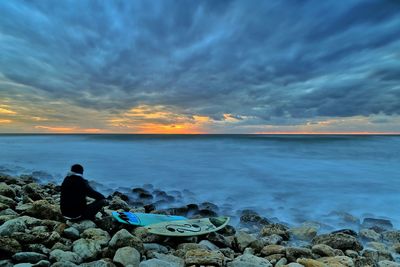 Man with surfboards on rocky sea shore against cloudy sky