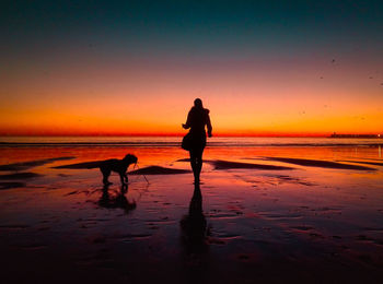 Silhouette woman with dog walking at beach against sky during sunset