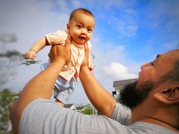 Low angle view of happy father carrying cute son while standing against blue sky