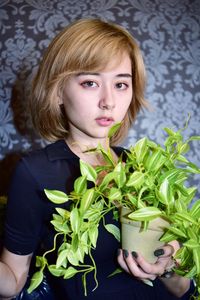 Portrait of beautiful woman holding plant standing against wall at home