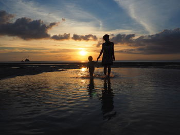 Silhouette mother and son holding hands standing on shore at beach against sky during sunset