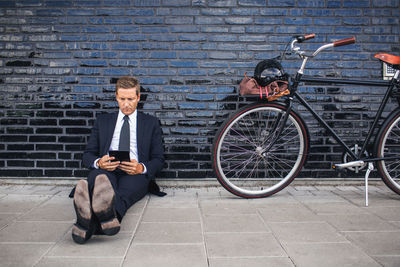Full length of businessman using digital tablet while sitting on sidewalk by bicycle