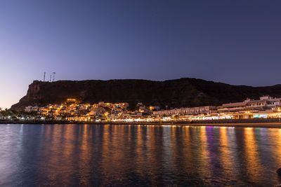 Illuminated buildings by sea against clear sky at night