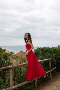 Woman wearing red dress while standing on railing against cloudy sky