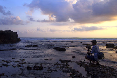 Man sitting on rock at beach against sky during sunset