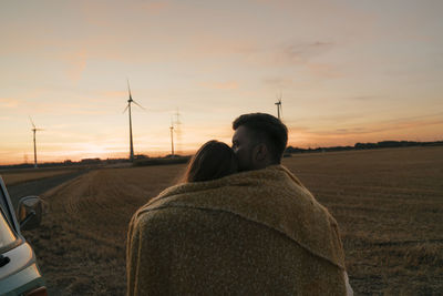 Couple wrapped in a blanket at camper van in rural landscape with wind turbines in background