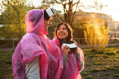 Happy young couple wearing raincoat and virtual reality simulator at park