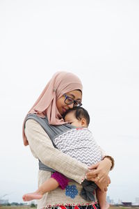 Mother wearing hijab carrying daughter against sky