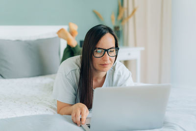 Brunette attractive young woman lying on the bed at home using her laptop