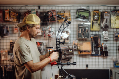 Side view of busy male hunter adjusting compound bow with arrow while standing in garage and preparing for shooting