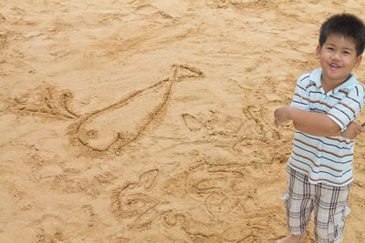 Portrait of smiling boy standing by fish drawing on sand at beach